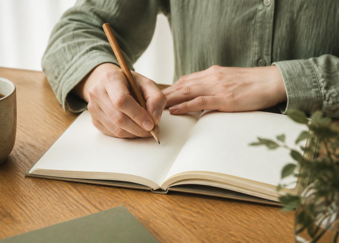 Hands writing notes in a lined notebook beside a stack of study materials.